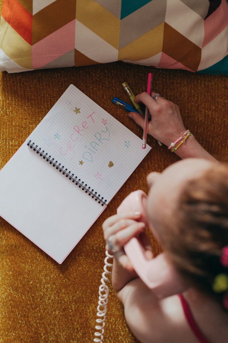 Overhead view of a woman writing in a diary with colored pens while talking on a pink telephone.