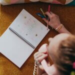 Overhead view of a woman writing in a diary with colored pens while talking on a pink telephone.